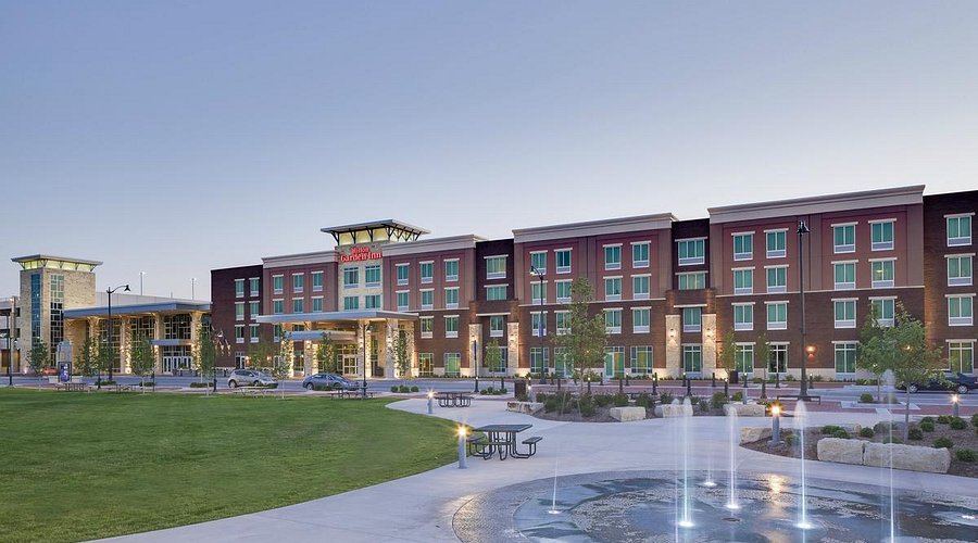 Exterior view of a Hilton Garden Inn hotel with four stories, modern architecture, and large windows. In front, there is a landscaped area with grass, trees, a paved walkway, benches, and a circular fountain with water jets. The sky is clear at dusk.
